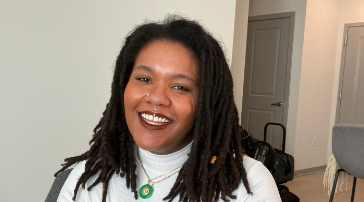 An African-American woman with dreads smiles. She is wearing a white long sleeve turtle neck and a plaid dress overtop.